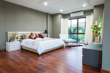 a large room with a
               hardwood floor, a tall window overlooking trees, a two-person bed with a single
               bedside table, and a padded chair next to the window